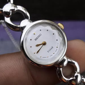 Könnte beinhalten: Eine silberne und goldene Seiko Tisse Uhr mit weißem Zifferblatt und schwarzen Zeigern. Die Uhr hat ein Gliederarmband.