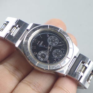 Könnte beinhalten: Eine silberfarbene Seiko-Armbanduhr mit schwarzem Zifferblatt. Die Uhr verfügt über drei Hilfszifferblätter und eine Wochentagsanzeige. Das Armband besteht aus silberfarbenem Metall. Ein klassisches Accessoire.