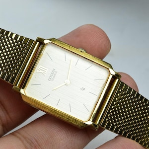 May include: A gold-tone rectangular Citizen Quartz wristwatch featuring a textured champagne-colored dial with thin hour markers, a Roman numeral at the twelve o'clock position, and a fine gold-tone mesh metal strap with a woven pattern.