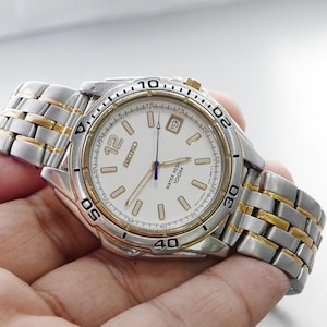 Könnte beinhalten: Silber-goldfarbene Seiko-Uhr mit weißem Zifferblatt, goldenen Akzenten auf Lünette und Armband. Die Uhr hat eine Datumsanzeige bei 3 Uhr und ist wasserdicht bis 100M.