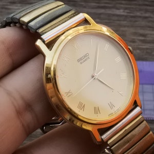 May include: A gold-tone Seiko quartz watch with a white face and Roman numeral hour markers. The watch has a two-tone metal band.