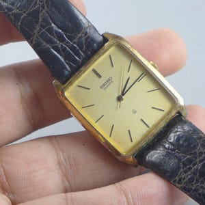 Puede incluir: Un reloj Seiko Quartz dorado con una esfera cuadrada y una esfera dorada. El reloj tiene manecillas y marcadores negros, y una correa de cuero negro. La esfera del reloj muestra el logotipo de Seiko.
