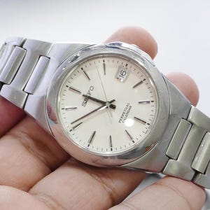 May include: A silver Seiko Perpetual Calendar watch with a white face and silver band. The watch face displays the date and the word "Seiko". The watch is held in a hand.