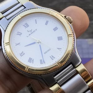 May include: A two-tone gold and silver watch with a white face and Roman numeral hour markers. The watch face also has the text "Landy" and "Seiko".