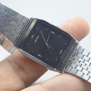 Puede incluir: Un reloj Seiko rectangular con esfera negra y correa plateada. La esfera del reloj tiene agujas e indicadores horarios plateados. La correa está hecha de pequeños eslabones rectangulares. El reloj está sujeto por una mano.