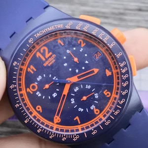 Puede incluir: Un reloj cronógrafo Swatch azul con detalles en naranja. El reloj tiene una esfera azul con números y agujas naranjas, una pantalla de fecha y una escala taquimétrica. Correa azul y botones naranjas.