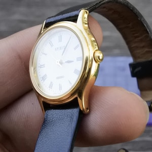 May include: A gold-tone wristwatch with a white face and black leather band. The watch face has Roman numeral hour markers and the word "Spirit" printed at the top.