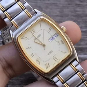 May include: A gold and silver two-tone Seiko Spirit watch with a square face and Roman numeral hour markers. The watch face is white with black hands and a date window that reads "TUE 13".