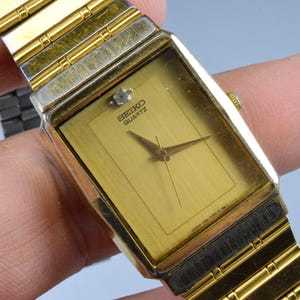 Puede incluir: Un reloj Seiko Quartz dorado con una esfera rectangular y una esfera dorada. El reloj tiene una correa dorada y un pequeño acento en forma de diamante encima del centro. Un reloj clásico.