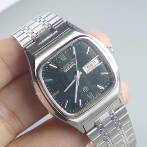 Pode incluir: Relógio Orient Quartz prateado com mostrador quadrado verde escuro. O relógio apresenta ponteiros e marcadores prateados, um visor de data e uma pulseira de elos prateada. O mostrador exibe o logotipo Orient Quartz.