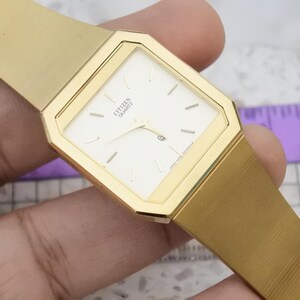 Könnte beinhalten: Goldfarbene Citizen Quartz-Uhr mit rechteckigem Zifferblatt und passendem goldenen Armband. Das Zifferblatt ist weiß mit schwarzen Markierungen und Zeigern. Klassisches Uhrendesign.