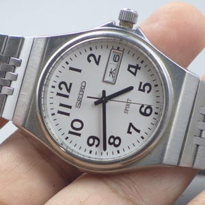 Könnte beinhalten: Eine silberfarbene Seiko Spirit Armbanduhr mit weißem Zifferblatt. Die Uhr hat schwarze Ziffern, Zeiger und eine Datumsanzeige. Das Armband besteht aus silberfarbenen Metallgliedern.