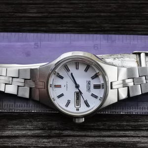 May include: A silver Seiko automatic wristwatch with a white face and a silver metal band. The watch face has black hour and minute hands, a red second hand, and a date window that displays "MON 29".