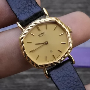 Könnte beinhalten: Eine goldfarbene Seiko Quartz-Uhr mit hellem Zifferblatt und schwarzem Lederarmband. Die Uhr hat eine achteckige Lünette und einfache Stundenmarkierungen. Das Zifferblatt zeigt das Seiko Quartz-Logo.