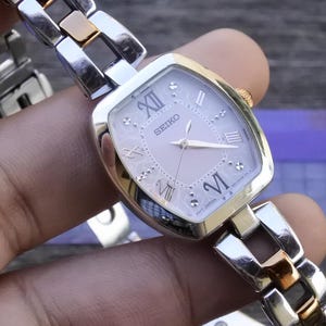 Könnte beinhalten: Eine silber- und goldfarbene Seiko Armbanduhr mit quadratischem Zifferblatt und römischen Ziffern. Die Uhr hat ein hellrosa Zifferblatt und ein Gliederarmband. Ein klassisches Accessoire.