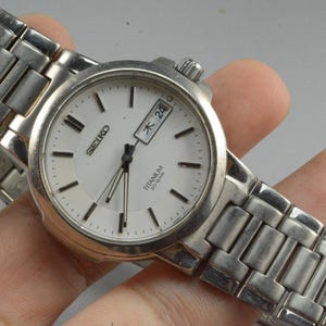 Könnte beinhalten: Eine silberfarbene Seiko Titanium Armbanduhr mit weißem Zifferblatt. Die Uhr hat eine Datumsanzeige und ein silbernes Gliederarmband. Die Uhr wird von einer Person gehalten.