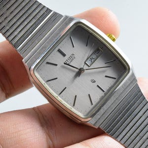Op de afbeelding: Een zilveren Citizen Quartz horloge met een rechthoekige wijzerplaat en een zilveren band. De wijzerplaat is lichtgrijs met zwarte uurmarkeringen en wijzers. Het horloge wordt door een hand vastgehouden.