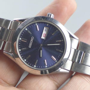 Pode incluir: Um relógio Seiko em aço inoxidável com mostrador azul. O relógio tem um visor de data e uma pulseira prateada. O relógio é segurado em uma mão.