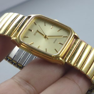 Könnte beinhalten: Goldfarbene Seiko Spirit Armbanduhr mit rechteckigem Zifferblatt und goldenem Armband. Das Zifferblatt hat eine hellgoldene Farbe mit schwarzen Stundenmarkierungen und Zeigern. Das Armband ist aus goldfarbenem, flexiblem Metall. Die Uhr wird von einer Hand gehalten.