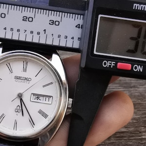 May include: A silver Seiko Quartz watch with a white face, black Roman numerals, and a date display. The watch is measured with a digital caliper, showing a diameter of 38.2 mm. The watch has a black leather strap.