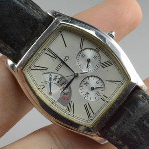 May include: A silver Seiko watch with a rectangular face and a black leather band. The watch face is light-colored with Roman numerals, three sub-dials, and the Seiko logo. The watch displays the day of the week and date.