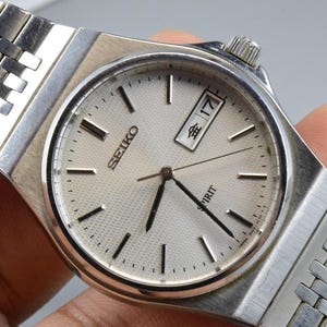 Könnte beinhalten: Eine silberfarbene Seiko Spirit Armbanduhr mit einem strukturierten cremeweißen Zifferblatt, silbernen Stabindizes, schwarzen Zeigern und einer Datumsanzeige auf der Drei-Uhr-Position mit dem Text 金 17. Sie hat ein Gliederarmband aus Edelstahl.