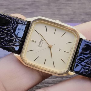 Puede incluir: Un reloj Citizen Quartz dorado con esfera cuadrada y correa de cuero negro. La esfera del reloj es de color crema con marcadores y agujas negras. La correa tiene un patrón texturizado similar al de un cocodrilo.