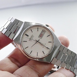 Op de afbeelding: Een vintage Seiko Quartz Type II horloge met een zilverkleurige metalen band en een witte wijzerplaat. De wijzerplaat toont de datum en tijdmarkeringen. Het horloge wordt in een hand gehouden.