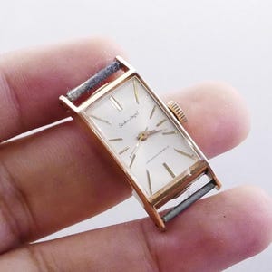 Puede incluir: Un reloj Seiko-Angel vintage en tono dorado con una esfera rectangular. El reloj tiene una esfera blanca con agujas e indicadores dorados. El reloj está en la mano de una persona.