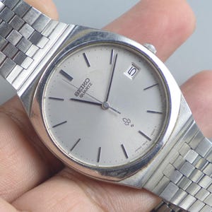 Op de afbeelding: Zilveren Seiko Quartz horloge met een rechthoekige wijzerplaat en een zilveren band. De wijzerplaat is lichtgrijs met zwarte uurmarkeringen en wijzers. De datum wordt weergegeven op 3 uur. De band is gemaakt van kleine, verbonden zilveren vierkantjes.