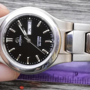 Könnte beinhalten: Eine silberne Citizen Eco-Drive Uhr mit schwarzem Zifferblatt und silbernen Zeigern. Die Uhr hat ein Datumsfenster bei 3 Uhr und die Wörter "Citizen Eco-Drive" und "Attesa" sind auf dem Zifferblatt aufgedruckt.