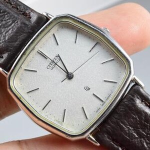 Pode incluir: Um relógio de pulso Citizen Quartz com caixa quadrada em tom prateado e mostrador prateado texturizado. O relógio apresenta marcadores de hora pretos finos, ponteiros pretos e uma janela de data na posição das três horas. Possui uma pulseira de couro castanho escuro.
