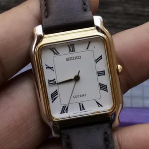 Puede incluir: Un reloj Seiko Lucent dorado con una esfera blanca y marcadores de hora romanos negros. El reloj tiene una correa de cuero negra.