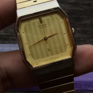Pode incluir: Um relógio de pulso Citizen Quartz dourado com um mostrador retangular e um mostrador dourado listrado. O relógio tem uma pulseira de metal dourado.