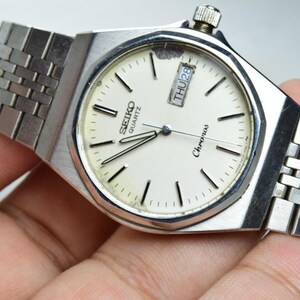 Könnte beinhalten: Eine silberfarbene Seiko Quartz Chronos Armbanduhr mit cremefarbenem Zifferblatt, silbernen Stabindizes und einer Datumsanzeige auf der Drei-Uhr-Position. Die Uhr verfügt über ein Edelstahl-Gliederarmband und eine metallische achteckige Lünette.