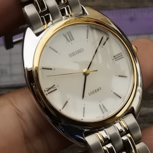 May include: A silver and gold two-tone Seiko Lucent watch with a white face and Roman numeral hour markers.