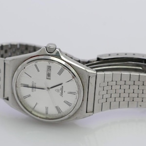 May include: A silver Seiko Quartz watch with a white face and a silver metal band. The watch face has black Roman numeral hour markers, black minute markers, and a date window at the 3 o'clock position. The date window displays the number 28 in Japanese characters.
