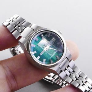 Puede incluir: Un reloj de pulsera vintage plateado con una esfera iridiscente verde y azul. El reloj tiene una correa de acero inoxidable con un diseño de eslabones clásico. El reloj se sostiene en una mano sobre un fondo claro.