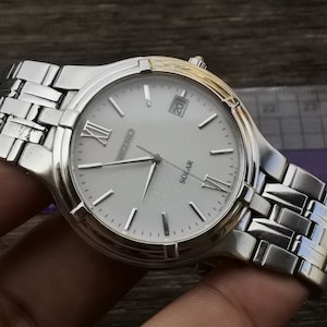 May include: A silver and gold men's wristwatch with a white face and black Roman numeral hour markers. The watch has a silver metal band and says "SEIKO SOLAR" on the face.