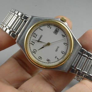 Könnte beinhalten: Eine silberfarbene Seiko Spirit Armbanduhr mit goldfarbener Lünette und weißem Zifferblatt. Die Uhr hat eine Datumsanzeige bei 3 Uhr und ein silberfarbenes Metallarmband. Auf dem Zifferblatt steht das Wort "spirit".