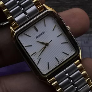 Könnte beinhalten: Eine Vintage-Session-Armbanduhr mit einem rechteckigen weißen Zifferblatt und goldfarbenen Akzenten. Die Uhr hat ein silbernes und goldfarbenes Armband. Das Wort "session" ist auf dem Zifferblatt aufgedruckt. Die Uhr wird in einer Hand gehalten.
