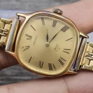 May include: A vintage gold-toned Seiko watch with a rectangular face and a gold-toned band. The watch face features Roman numerals and the words "Seiko" and "Jocie 24 Jewels". The watch is a classic timepiece.