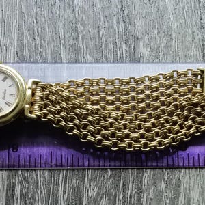 May include: A gold-tone wristwatch with a white face and a gold mesh band. The watch face has black Roman numeral hour markers and black hands. The watch band is made of a series of interlocking gold links.