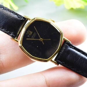 Könnte beinhalten: Eine Vintage Seiko-Uhr mit schwarzem Zifferblatt und goldfarbenem Gehäuse. Die Uhr hat ein schwarzes Lederarmband und goldene Zeiger. Das Zifferblatt hat römische Ziffern. Eine klassische Uhr.