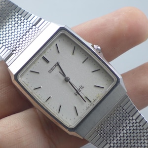 Puede incluir: Un reloj de pulsera Seiko Spirit plateado con esfera cuadrada y correa de malla. El reloj tiene una esfera de color claro con marcadores y agujas negras. El reloj lo sostiene una persona.