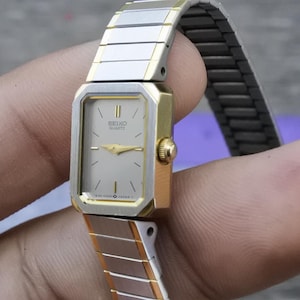 Puede incluir: Un reloj de dos tonos, plateado y dorado, con una esfera cuadrada y una correa plateada. La esfera es blanca con agujas negras y marcadores de hora romanos negros. La esfera también tiene el texto "SEIKO Quartz" impreso en ella.