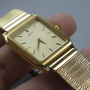 Puede incluir: Un reloj Seiko dorado con una esfera rectangular y una correa dorada texturizada. La esfera del reloj es de color oro claro con marcadores negros y agujas doradas. La correa es de estilo malla con un patrón repetido.