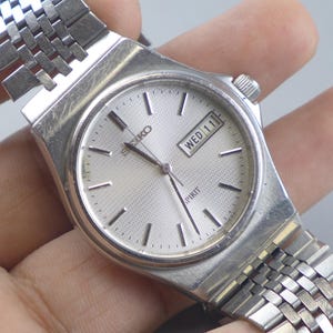 Könnte beinhalten: Eine silberne Seiko Spirit Armbanduhr mit strukturiertem Zifferblatt und Datumsanzeige "MIT 11". Die Uhr hat ein silbernes Armband und ein rechteckiges Gehäuse. Die Uhr ist ein klassischer Zeitmesser.