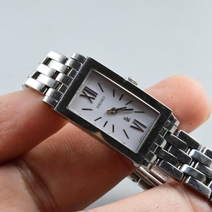 Könnte beinhalten: Eine rechteckige Seiko-Armbanduhr mit silberfarbenem Metallarmband und Gehäuse. Das Zifferblatt ist weiß mit schwarzen römischen Ziffern und schwarzen Zeigern. Die Uhr wird in einer Hand gehalten.