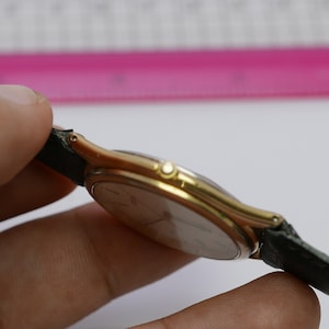 Könnte beinhalten: Eine goldfarbene Armbanduhr mit einem schwarzen Lederarmband. Das Zifferblatt der Uhr ist rund und hat ein silbernes Zifferblatt.